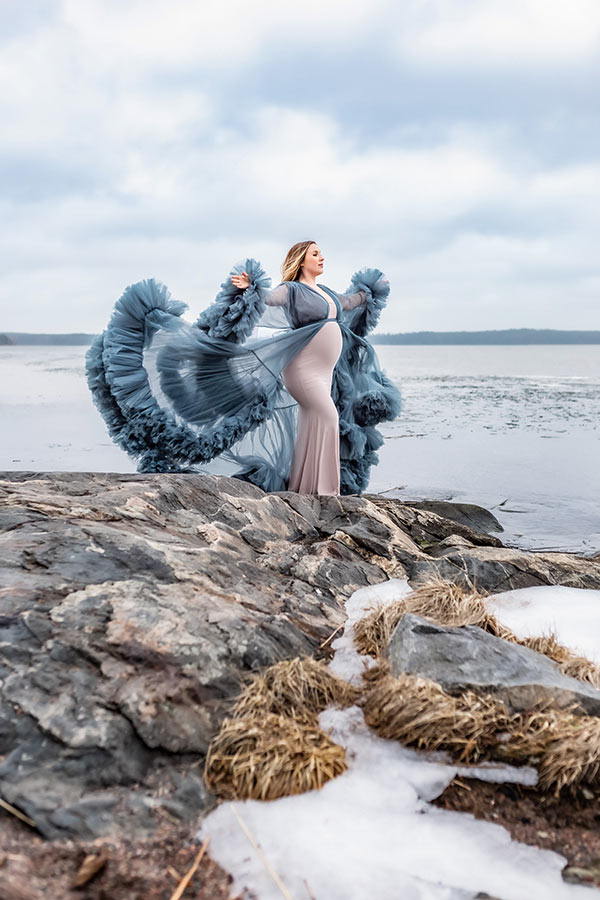 Gravidfotografering vid en klippa som delvis täcks av is. I bakgrunden syns ett isigt hav och den gravida kvinnan sträcker ut armarna medan den blå klänningen fladdrar i vinden.