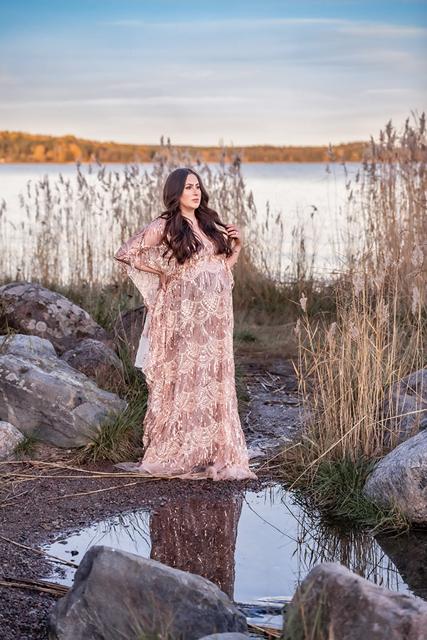 En gravid kvinna i glittrande gravidklänning står på en liten strand bland höga vass och stora stenar. I bakgrunden skymtar havet och i förgrunden syns ett stilla vatten med kvinnans reflektion.