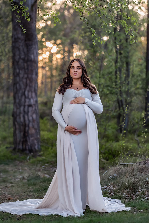 Gravid kvinna står i skogen med vackert solljus. På sig har hon en beige gravidklänning med släp.