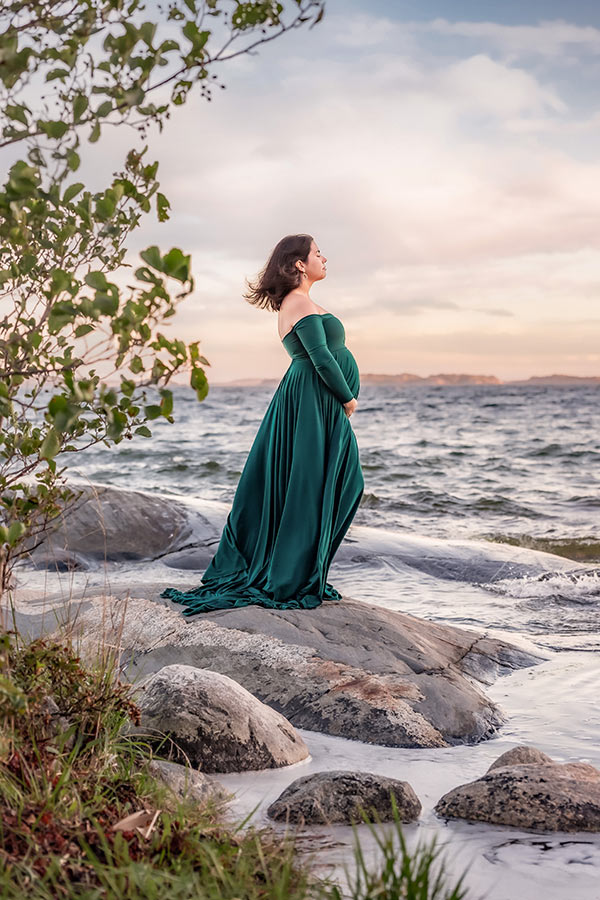 Gravidfotografering i skärgården vid havet. Den gravida kvinnan har en grön klänning och står på klipporna där vågorna slår. I förgrunden syns grenar med löv och gräs och himlen är färgad av kvällsljuset.
