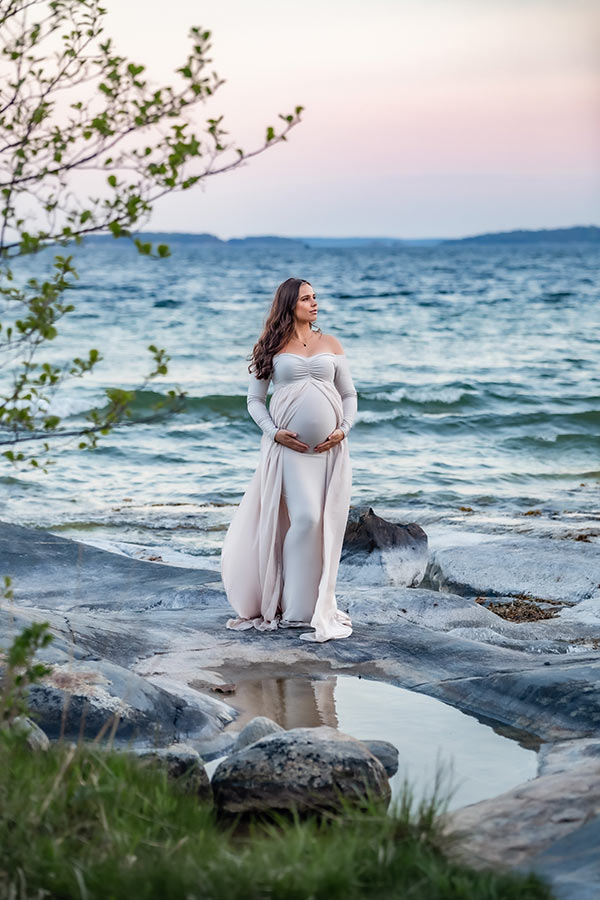 Gravidfotografering vid havet. Vågorna går höga i bakgrunden och i förgrunden är ett stilla vatten i klipporna. Den gravida kvinnan har en elegant beige gravidklänning med släp.