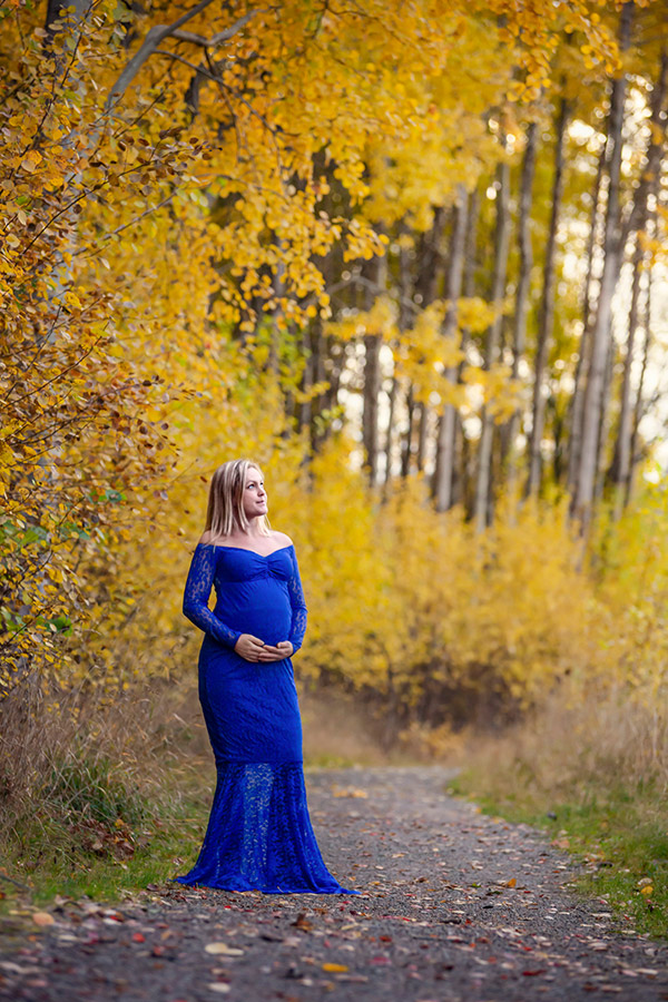 Gravidfotografering i oktober där den gravida kvinnan står på en grusgång omgiven av gula träd. Kvinnan har på sig en blå spetsklänning och på grusgången ligger löv.