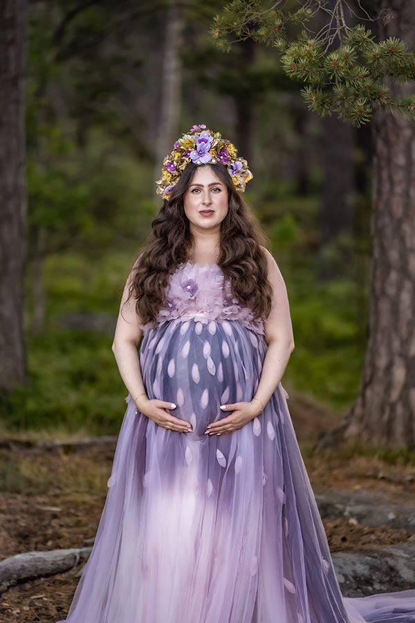 Gravidfotografering i en barrskog där den gravida kvinnan har en lila tyllklänning med blommor och blad samt en blomstertiara.