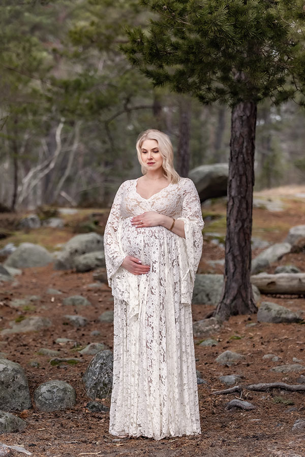 En gravid kvinna i vit spetsklänningar står i en barrskog och tittar ner på magen.