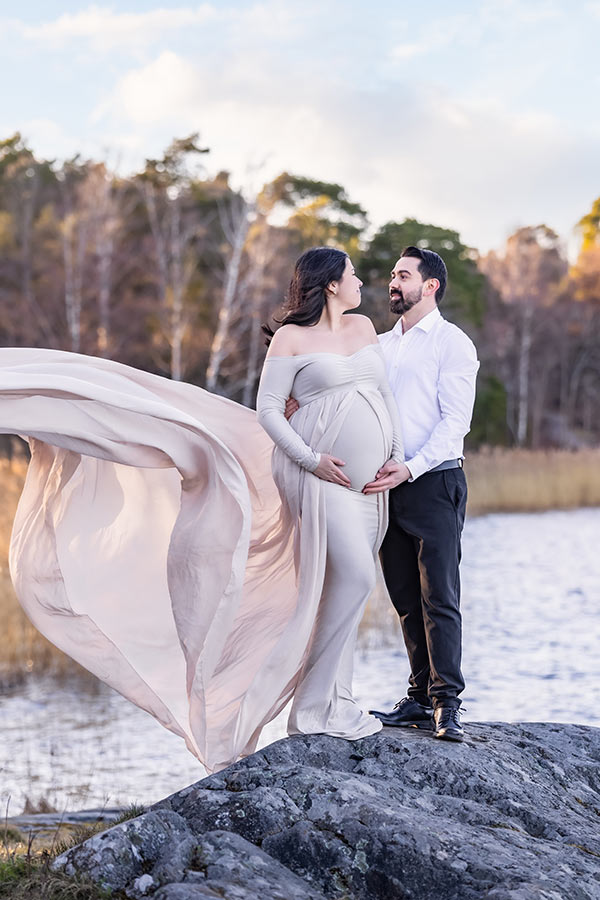 En gravid kvinna och hennes man står på en klippa vid havet. I bakgrunden syns träd och vass. De tittar på varandra och kvinnans klänningssläp flyger i vinden.