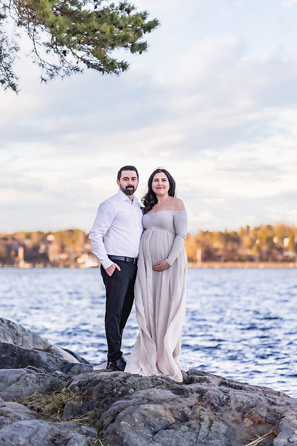 Ett vackert par står på en klippa vid havet under en tallgren och tittar in i kameran. Kvinnan är gravid och paret är stiligt klädda i beige, svart och vitt.