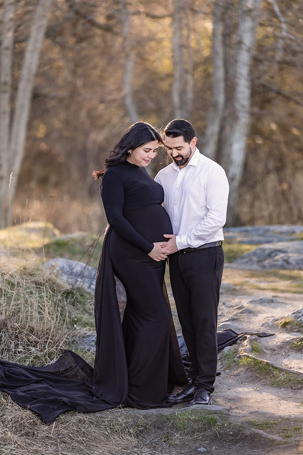 Gravidfotografering på en solupplyst glänta. Paret tittar ner på magen och ler.
