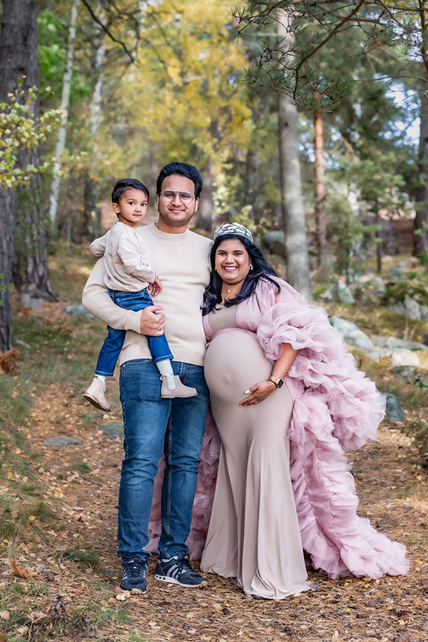 En familj, bestående av en gravid mamma, en pappa och en liten blivande storebror, står glada i en höstig skog.