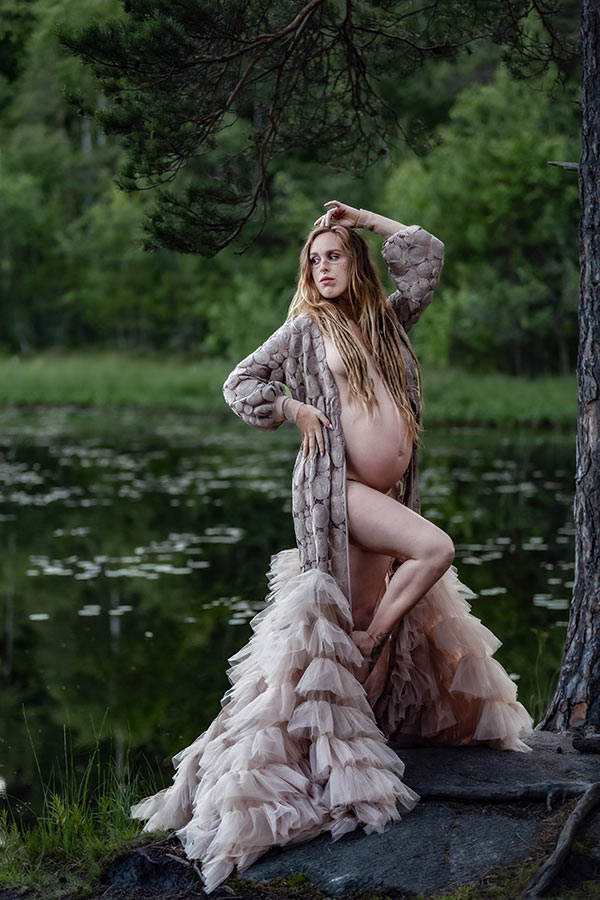 Gravidfotografering vid en tjärn i en skog. Kvinnan står på klippor vid vattnet och har på sig en beige fantastisk gravidklänning som är öppen och visar magen.