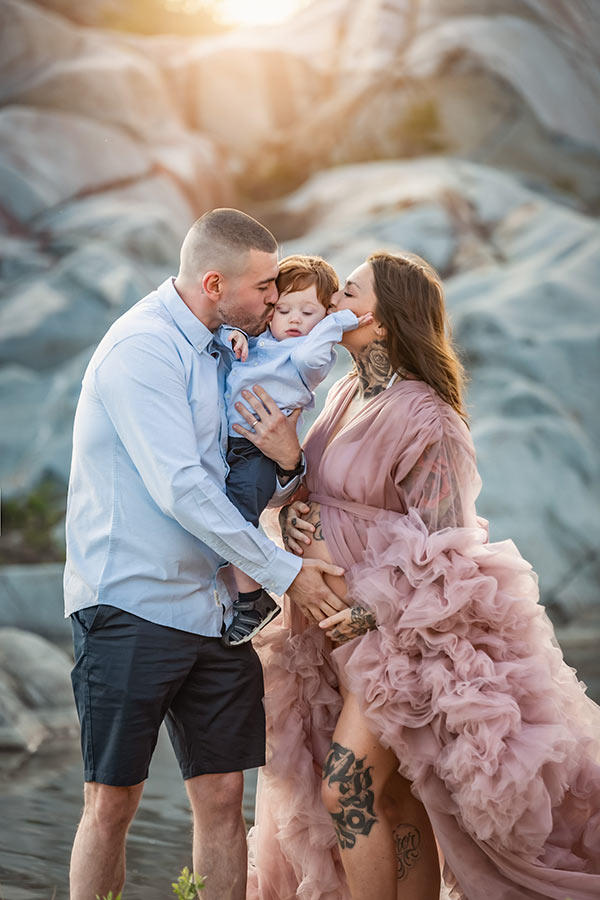 En kärleksfull familj fotograferas i ett klipplandskap. Mamman, som är gravid, och pappan håller upp och pussar deras ettåriga son.
