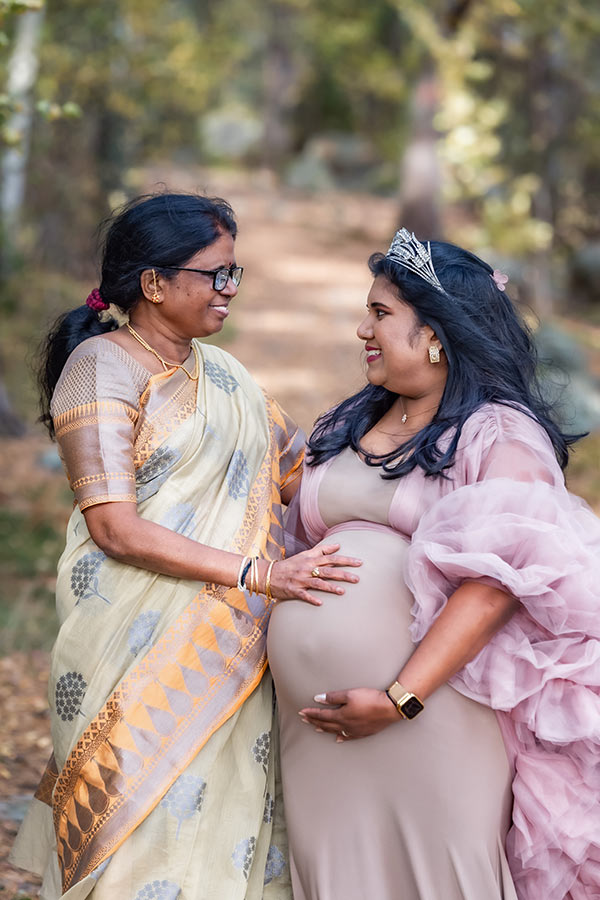 En gravid kvinna, med tyllklänning och silverkrona, står tillsammans med sin mamma, som har på sig vackra indiska kläder, står på en skogsstig. Den blivande mormodern håller handen på sin dotters mage.