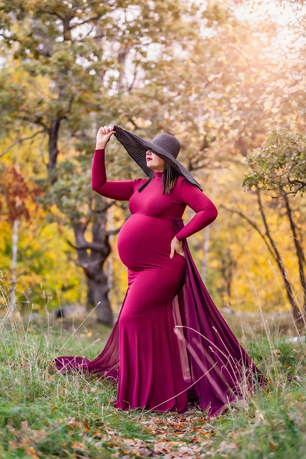 Gravidfotografering i oktober bland höstträd och höga gräs. Den gravida kvinnan har på sig en röd klänning med långt släp och en svart hatt med gigantiskt brätte.