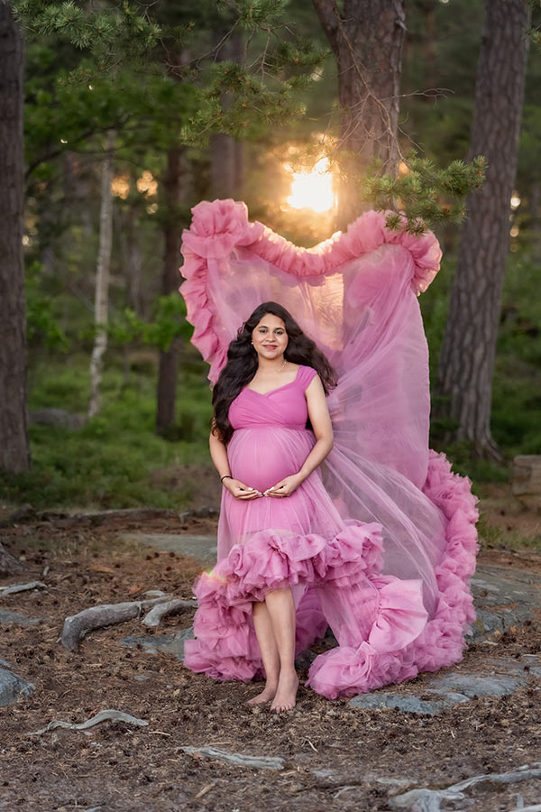 En kvinna med ett långt tyllsläp på sin rosa klänning står i en barrskog. Solen skiner bakom kvinnan som är gravid och tyllsläpet flyger i luften.