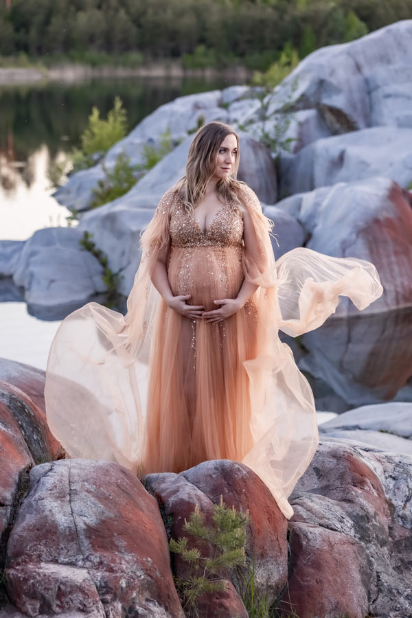Gravidfotografering på klipporna vid en liten sjö. Den gravida kvinnan har en persikofärgad lyxig gravidklänning som flyger i vinden.