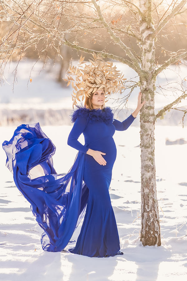 Gravidfotografering på vintern i ett snötäckt landskap. Den gravida kvinnan står vid en björk och har på sig en blå gravidklänning och en stor guldig krona av guldblad. Solen skiner starkt i bakgrunden.
