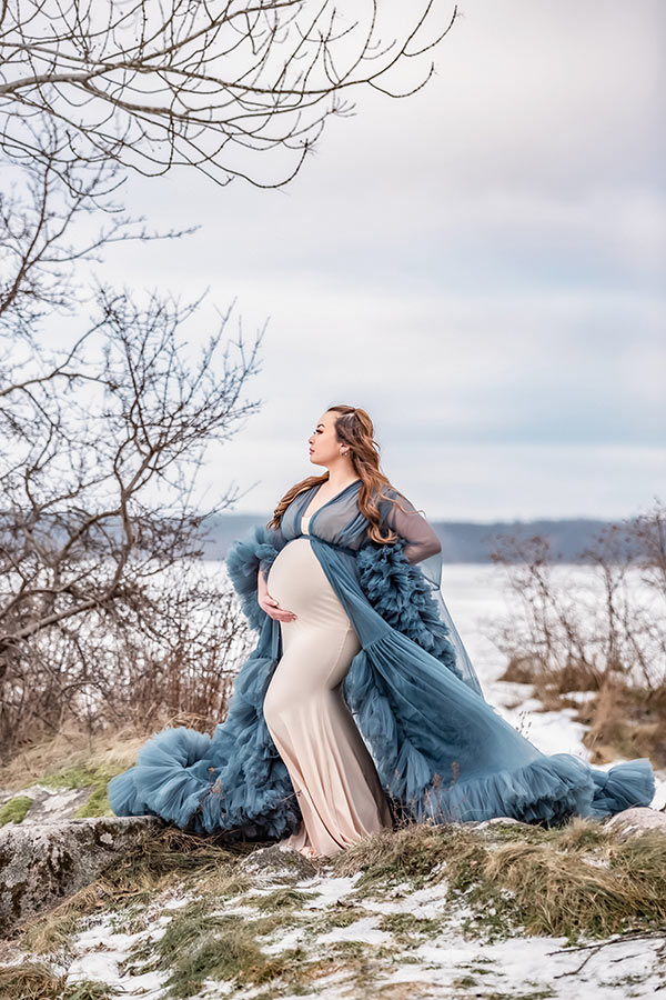 Gravidfotografering under vintern. Den gravida kvinnan står bland växtlighet och i bakgrunden skymtar ett fruset hav.