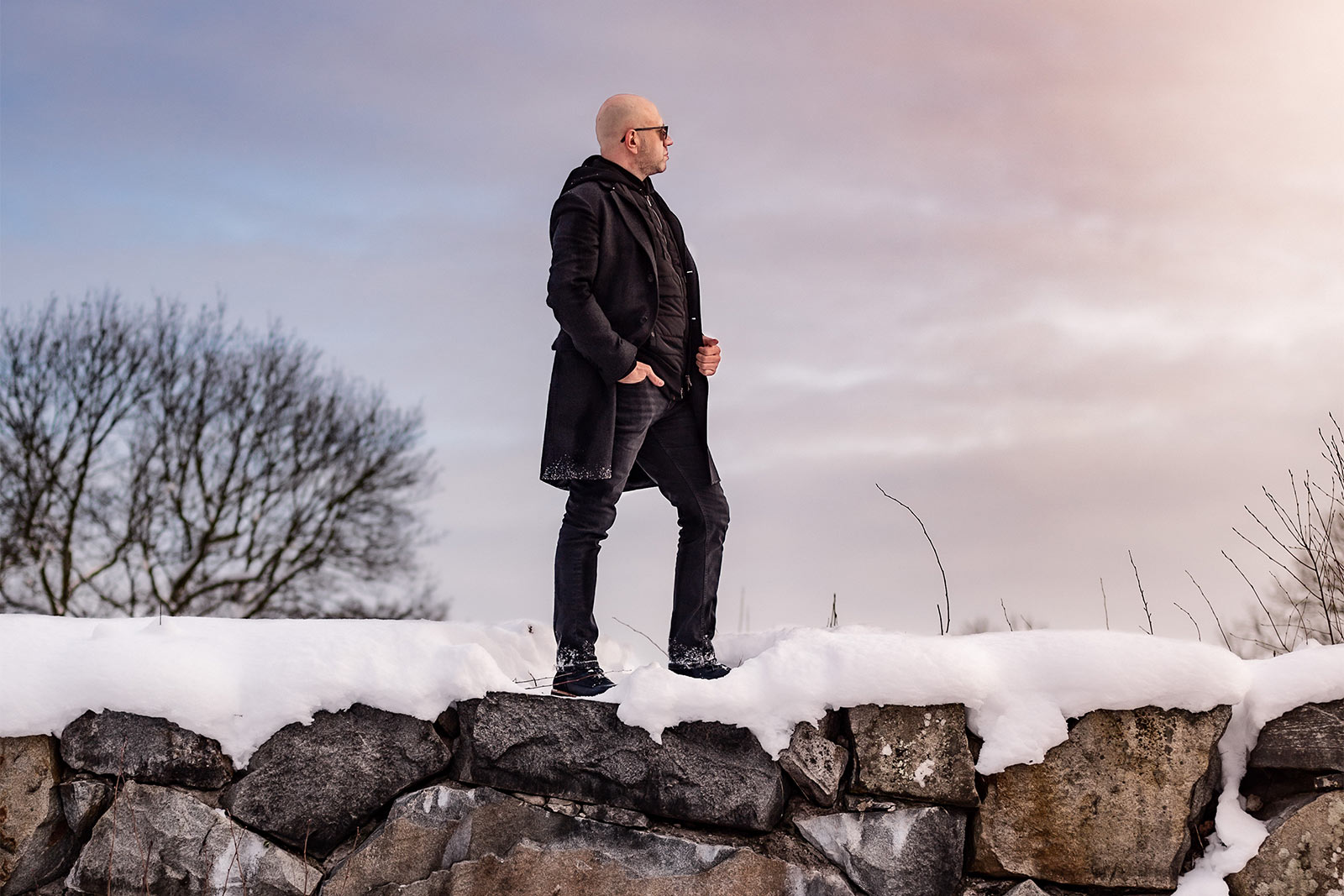 Porträttfotografering en snörik och kall vinterdag. Mannen som fotograferas är svartklädd och står uppe på en snötäckt stenmur. I bakgrunden är himmelen rosa.