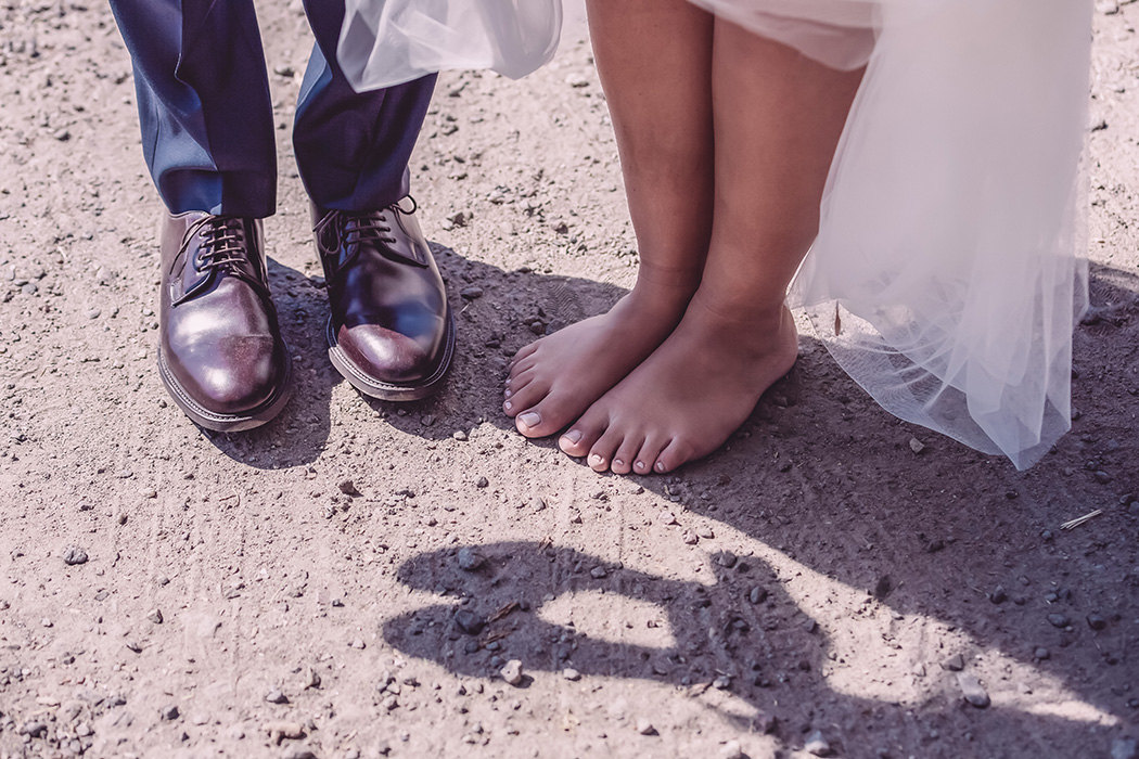 Två par fötter står på en grusgång, det ena paret har på sig manliga finskor medan det andra paret är barfota. I skuggan som faller på grusgången syns det att någon håller i ett par högklackade skor.
