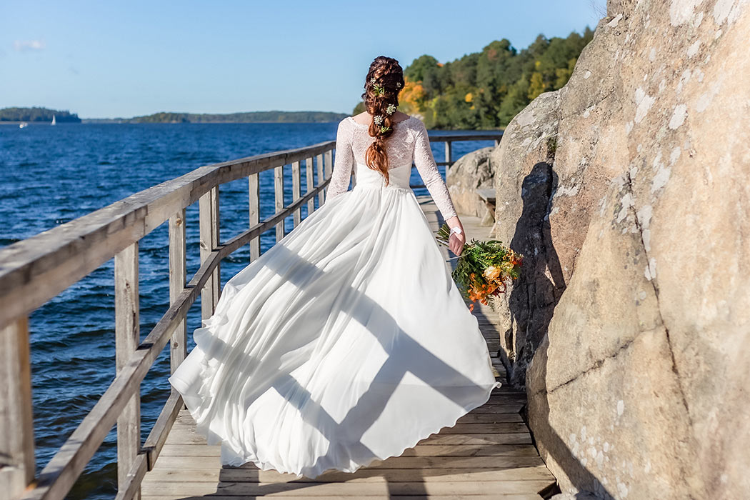 En brud klädd i bröllopsklänning och bärandes en brudbukett går på en bryggpromenad. Brudklänningen fladdrar i vinden och i bakgrunden syns vatten med segelbåt och höstiga träd.