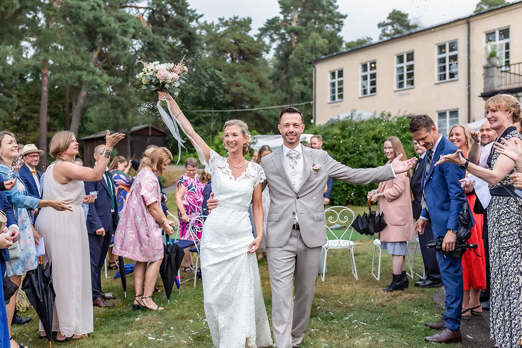 Det glada nygifta paret går mot bröllopsfotografens kamera medan gästerna runt omkring kastar ris och hyllar kärleken. De har precis gift sig i Mästervillans trädgård på Lidingö.