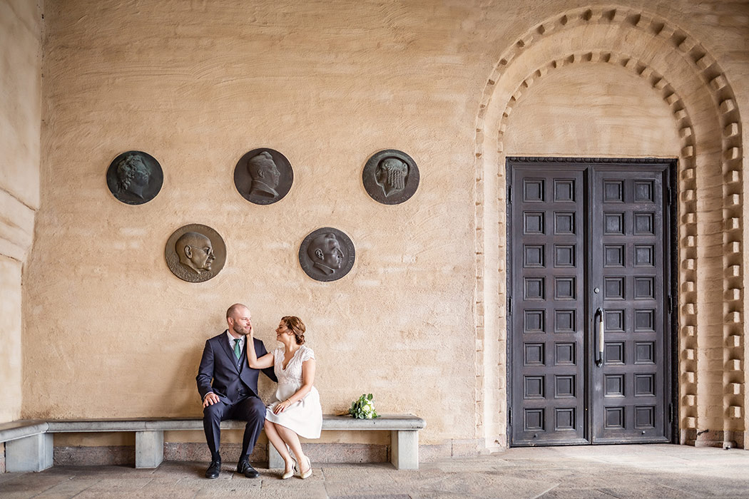 Brudpar sitter på en stenbänk vid en stor pampig dörr. Bruden klappar brudgummen på kinden och bredvid paret ligger brudbuketten.