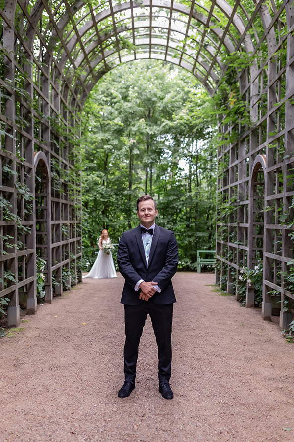 Fotografering av first look. Brudgrummen står i ett vackert uterum med grönska runt och väntar på bruden som kommer gående i bakgrunden.