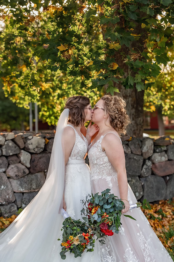 Bröllopsfotografering i Gamla Uppsala under ett stort höstfärgat lönnträd. Brudarna har vackra vita brudklänningar och kysser varandra. De har fantastiska brudbuketter i höstfärger.