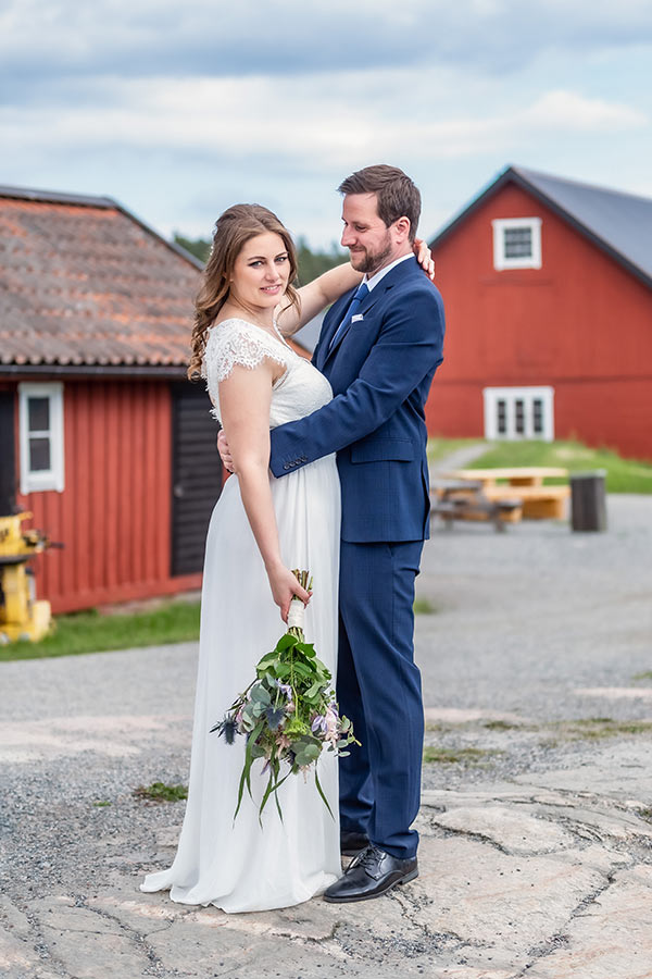 Bröllopsfotograf fotograferar bröllopspar i grönskande natur i Rönninge By i Täby. Det regnar så brudparet kramas under ett paraply och tittar in i kameran.