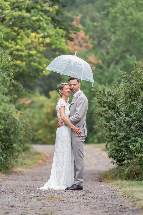 Bröllopsfotograf fotograferar bröllopspar i grönskande natur. Det regnar så brudparet kramas under ett paraply och tittar in i kameran.