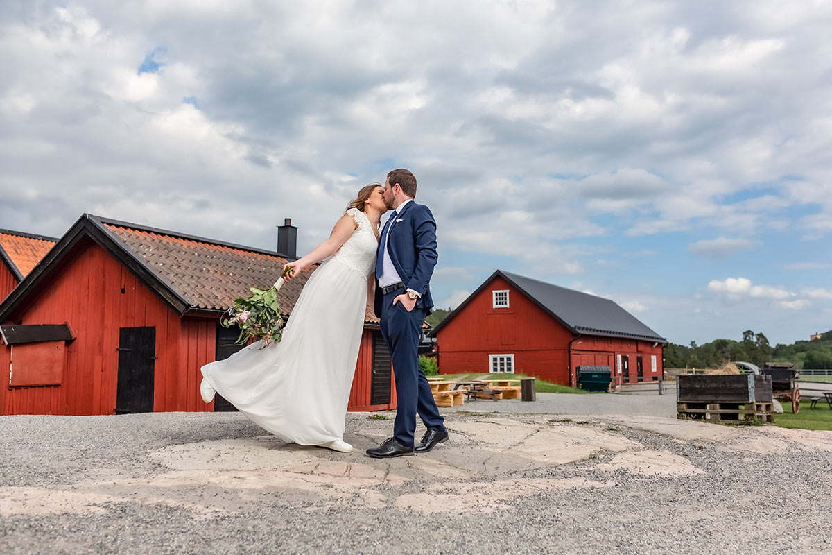 Ett nygift brudpar kysser varandra bland röda ladugårdar i Rönninge By i Täby.