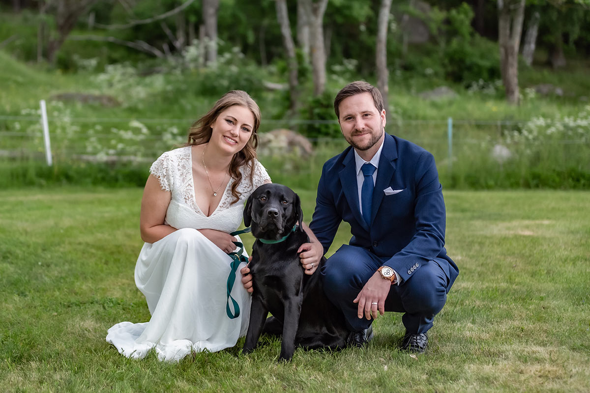Ett nygift par fotograferas sittandes vid sin hund på en äng.