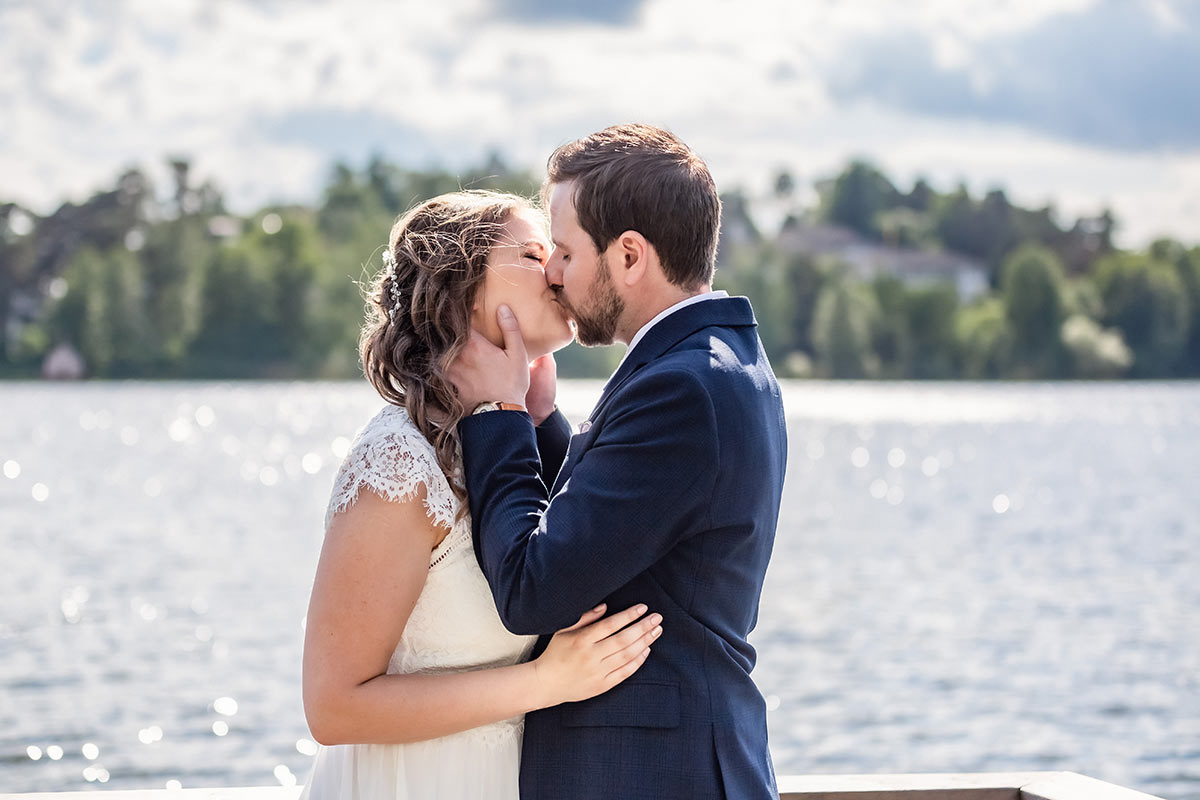 En brud och brudgrum kysser varandra vid en glittrande somrig sjö.