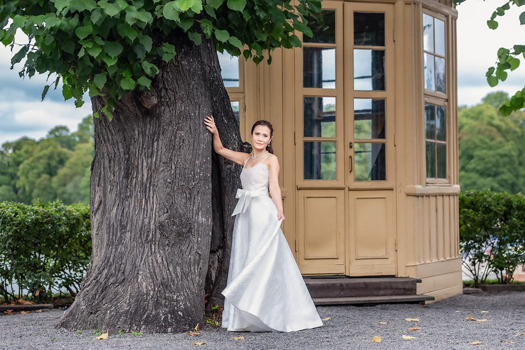 En brud i vacker klänning står vid ett stort träd vid ett lusthus. Hon tittar in i kameran och svänger med kjoldelen.