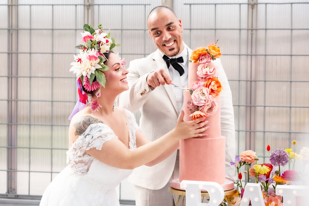 En brud håller i en tårta medan brudgummen skär tårtan. Tårtan är rosa och dekorerad med rosa blommor. Brudparet skrattar.
