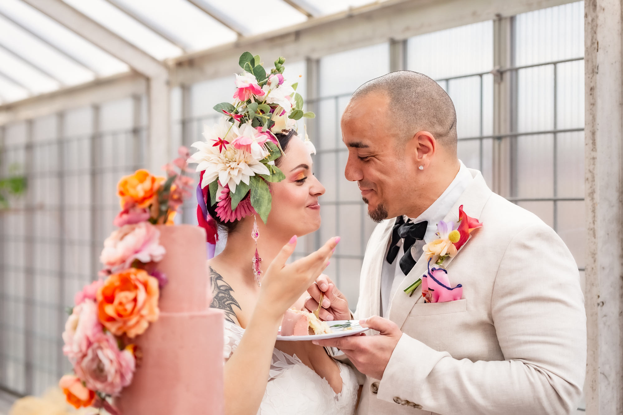 Bruden och brudgummen äter en tårtbit tillsammans. De tittar på varandra och ler. I förgrunden syns bröllopstårtan som är rosa med levande blommor.