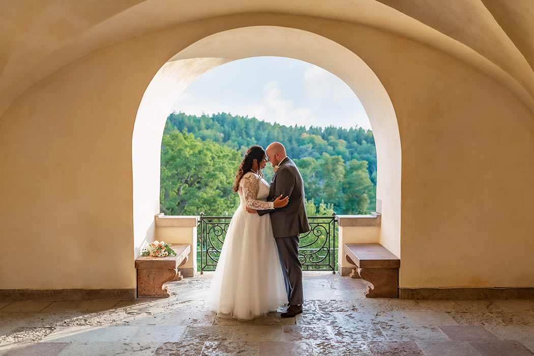 Bröllopspar håller om varandra framför en öppning i en slottsmur. Utsikten visar trädtoppar och en blå himmel.
