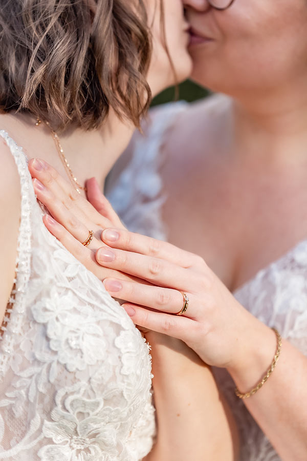 Fokus på händer med vigselringar medan brudparet pussas i bakgrunden.