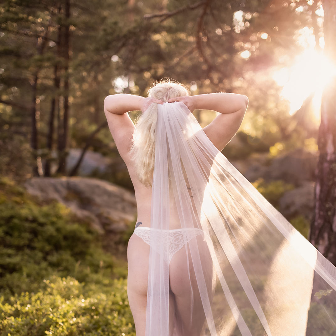 Boudoirfotografering i skogen i solnedgången. Kvinnan har vita underkläder och en vit slöja.