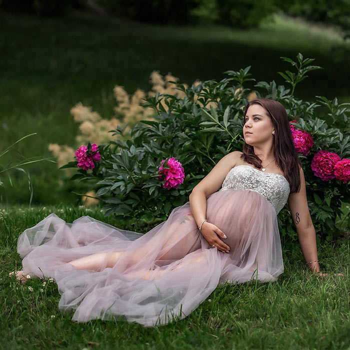 Gravidfotografering på sommaren. Den gravida kvinnan sitter framför en blommande pionbuske.