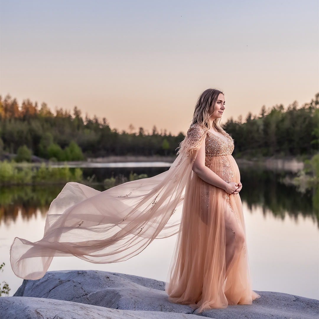 En gravid kvinna står på klippor vid ett spegelbankt vatten, hennes klänning glittrar och flyger i vinden.