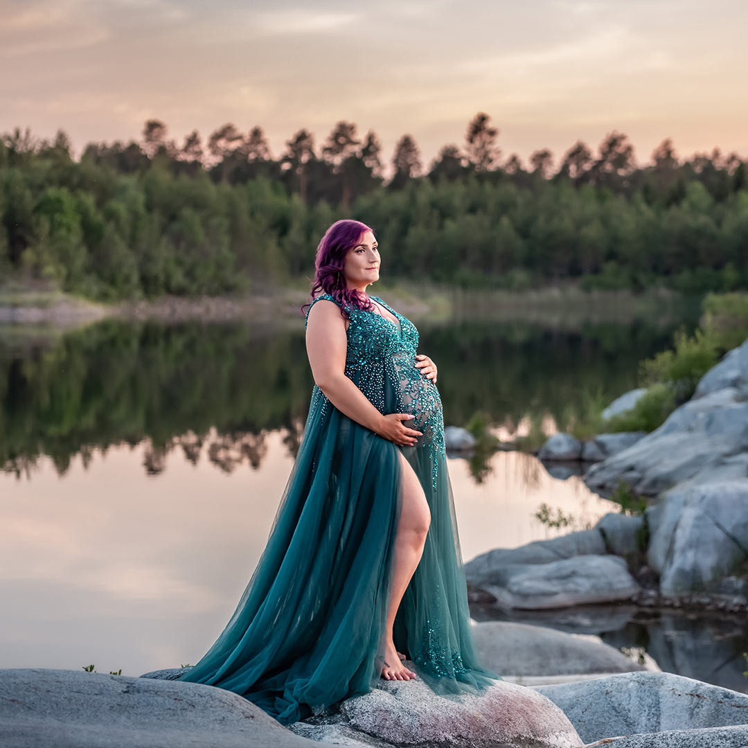 Gravidfotografering i ett stenbrott i det mjuka kvällsljuset. På klipporna står den vackra kvinnan i en grön och glittrig gravidklänning och på andra sidan vattnet speglas en grön skog.