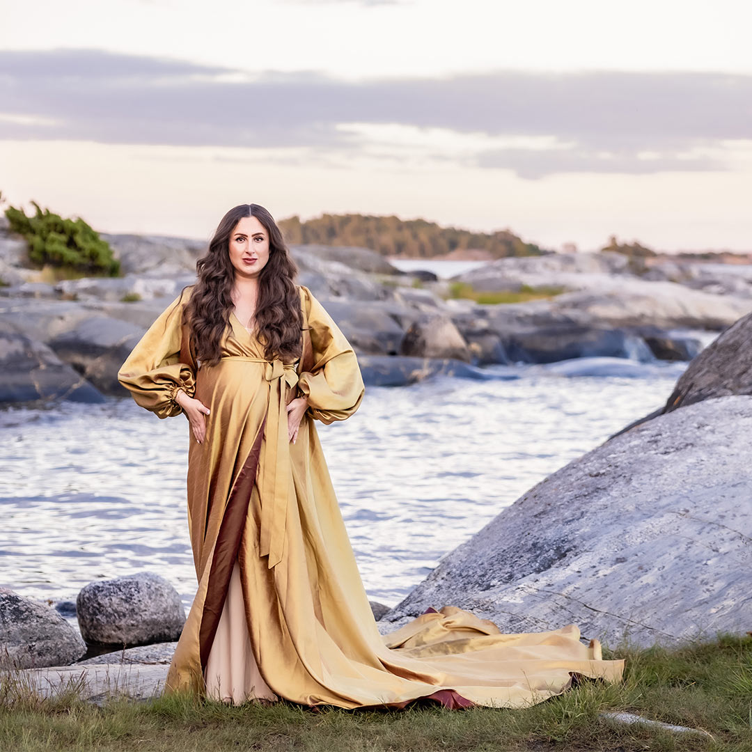 En gravid kvinna i extravagant guldklänning står bland klipporna i skärgården.