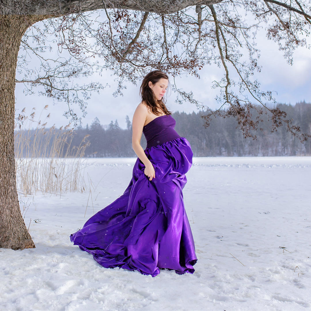 En gravid kvinna i lila satinklänning går över den snöiga isen bredvid ett stort träd.