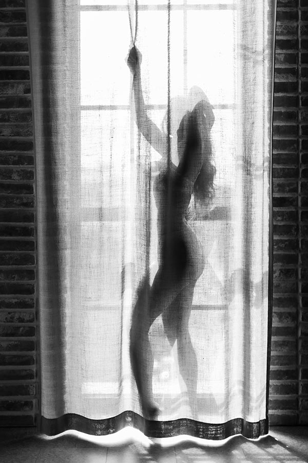 En kvinna står bakom en genomskinlig gardin. Solen skiner in från fönstret och kvinnan syns som en siluett mot gardintyget.