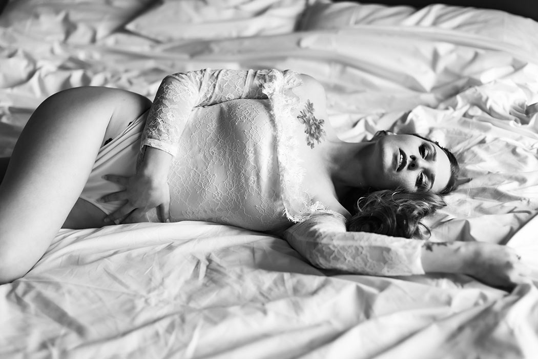 Svartvit boudoirbild där den vackra kvinnan ligger i en säng och har en vit spetsbody.