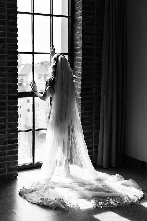 En vackert stilistisk bild på en kvinna som stor och tittar ut genom ett stort fönster som går från golv till tak. Bilden är svartvit och kvinnan har en lång slöja som får ett vackert mönster av det starka solljuset.
