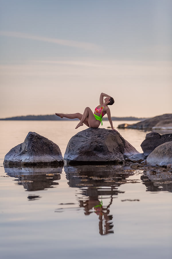 En kvinna sitter på en sten ute i vattnet iklädd en neonfärgad body. Hennes reflektion speglas i vattnet.