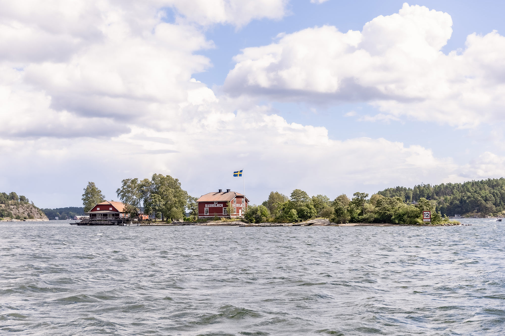 Stockholms skärgårdsö Badholmen utanför Vaxholm. Bilden är tagen på en båt på väg från Vaxholm till Badholmen. På ön står två röda stugor med vita knutar, på det ena huset står det Badholmen med stora vita bokstäver.