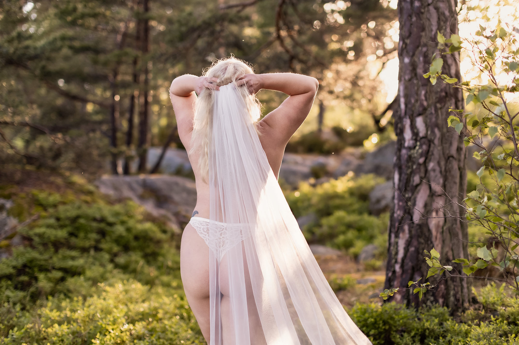 Boudoirfotografering i en skog i kvällsljus. Kvinnan syns bakifrån och har på sig en vit slöja och vita underkläder.