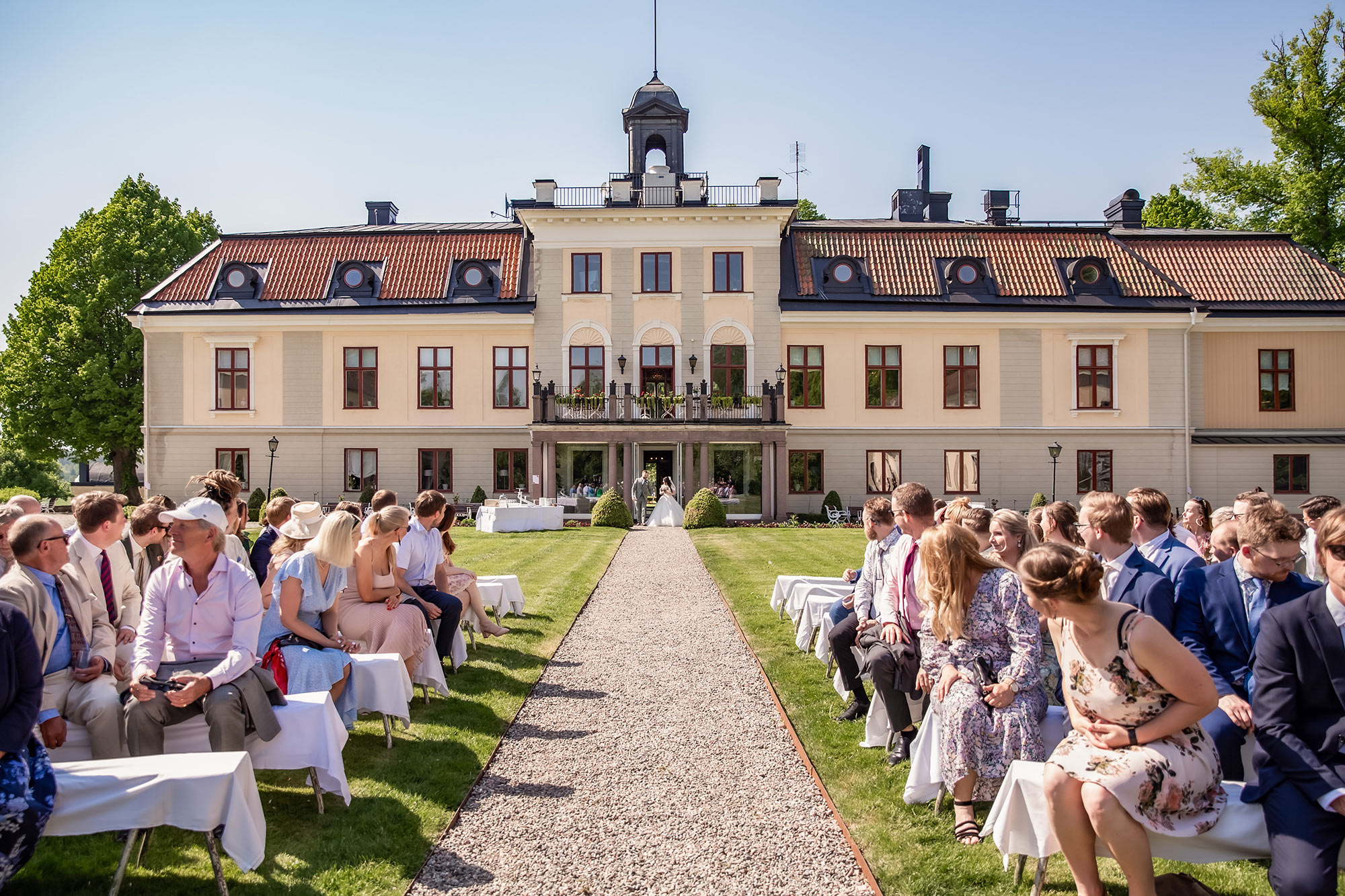 Bröllop på Södertuna Slott i deras slottspark. Gästerna sitter på bänkar och brudparet kommer precis ut från slottet och går mot vigselplatsen.